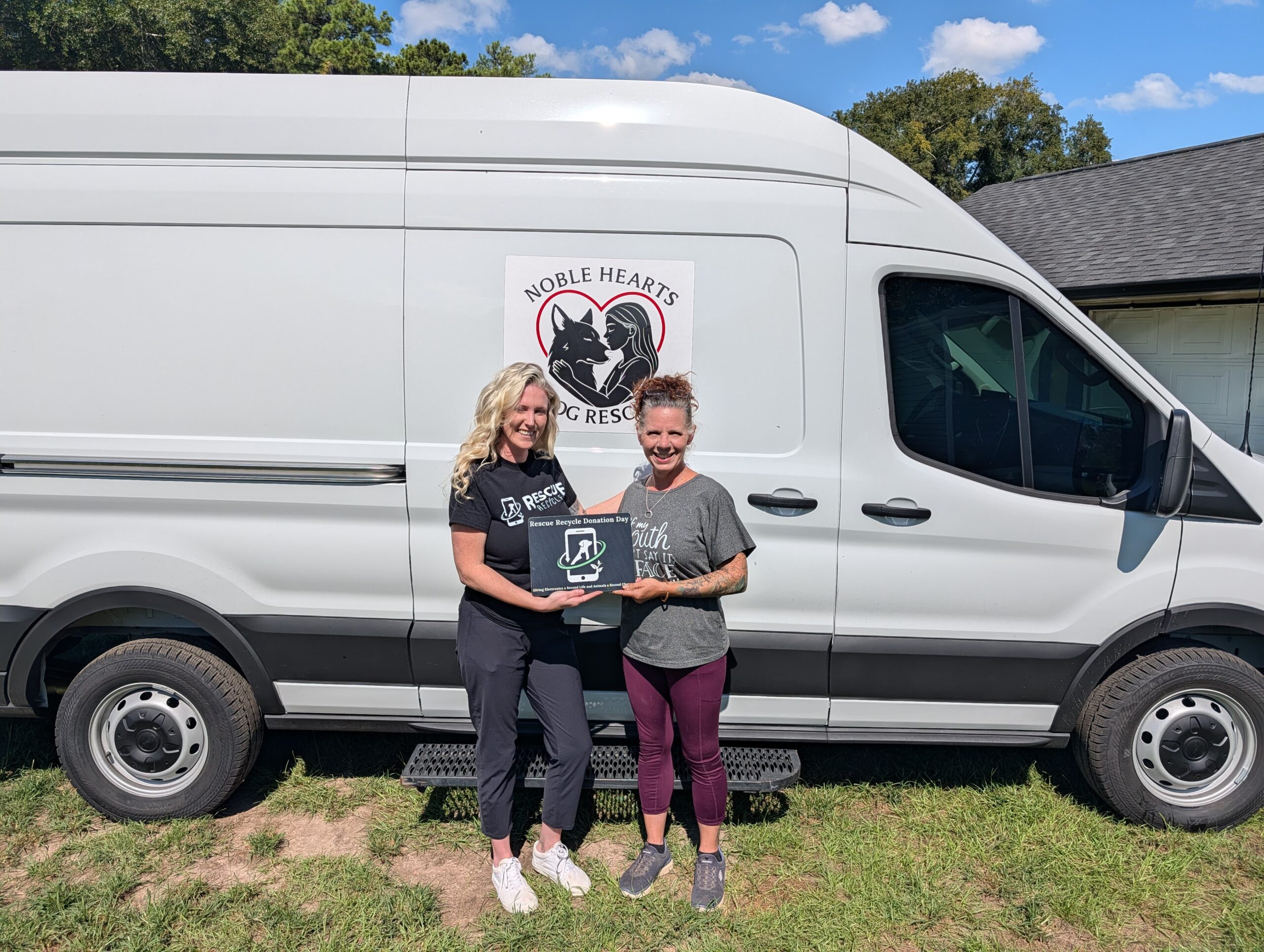 Two women holding a Rescue Recycle Donation Day sign in front of a van with the Noble Hearts Dog Rescue logo